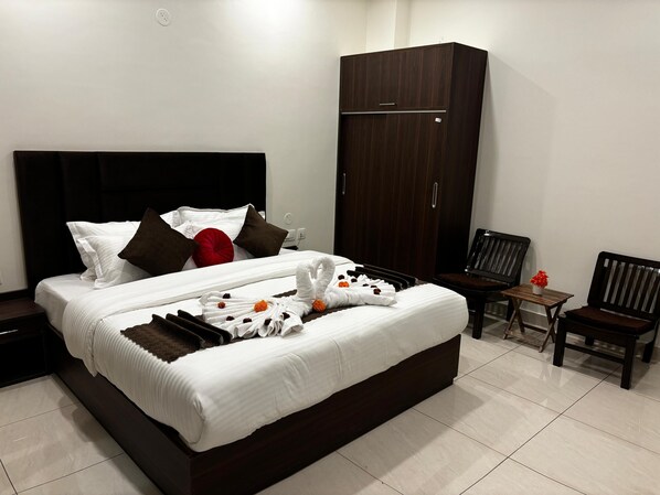 A neatly made bed with white linens, a dark headboard, and a red pillow. There is a wooden wardrobe to the right and two wooden chairs with a small table beside the bed.