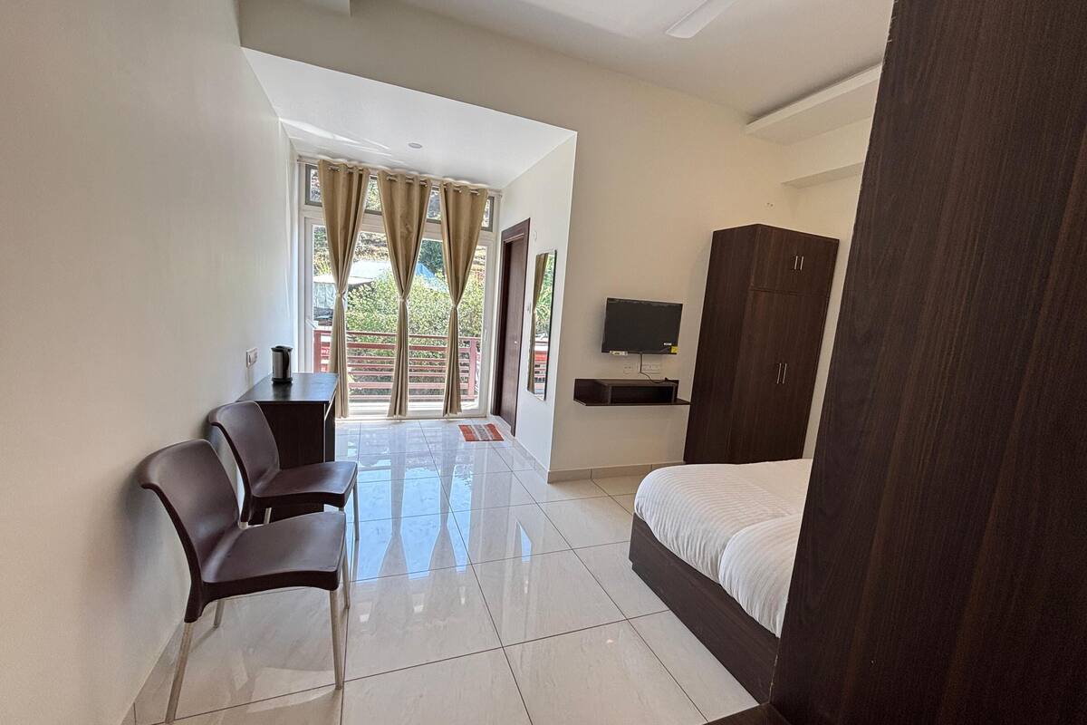 A hotel room with a bed, a desk with chairs, a TV mounted on the wall, and a wardrobe.