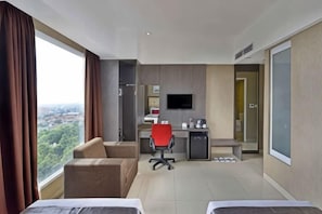 A hotel room with a bed, a desk with a chair, a TV, and a view of the city. - Travello Hotel Bandung (Cidadap)