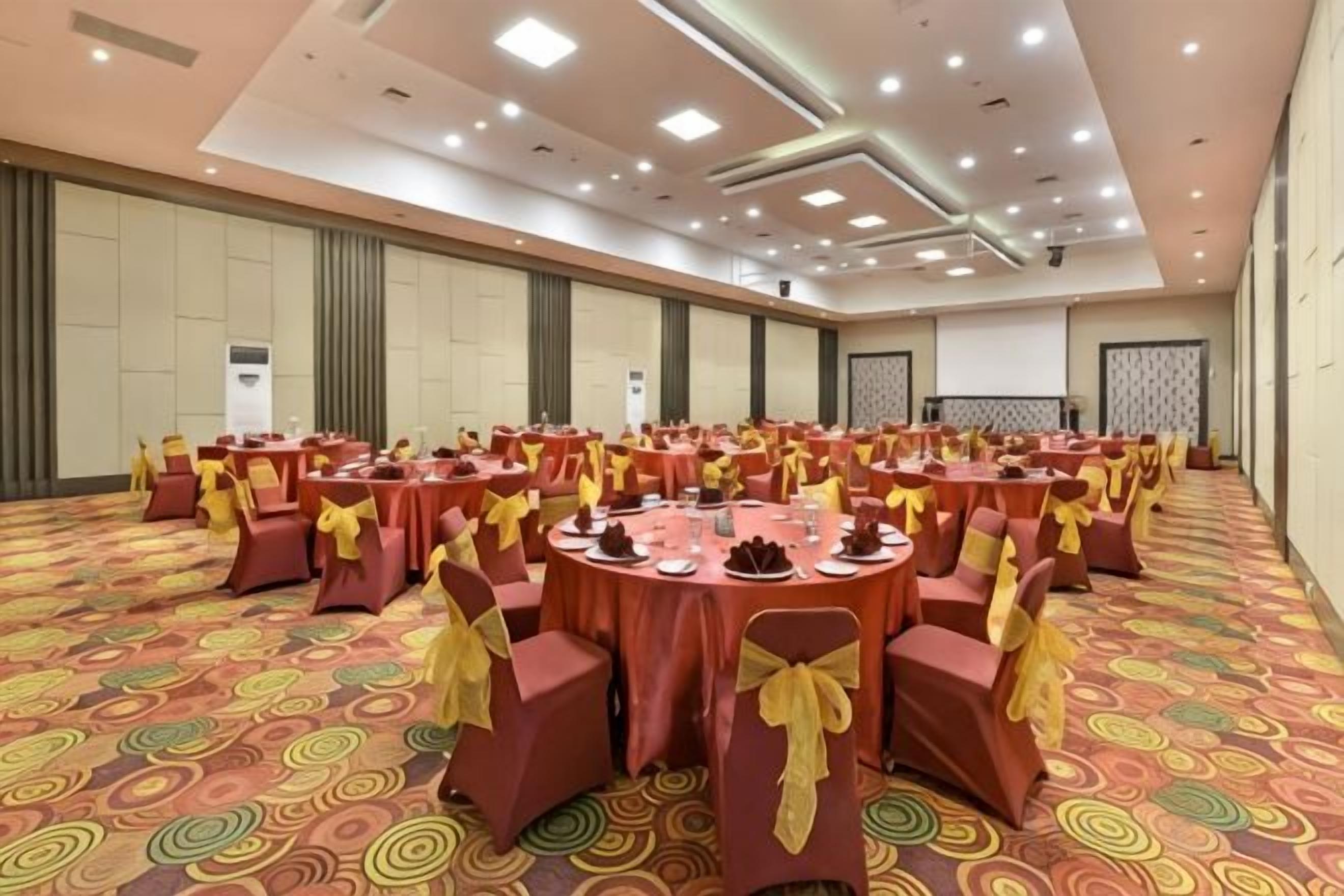 A large banquet hall with round tables and chairs, carpeted floor, and a high ceiling with recessed lighting.