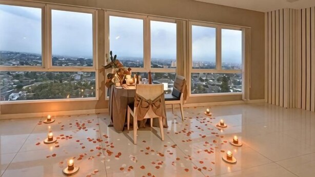 Ein Zimmer mit großem Fenster und Blick auf eine Stadtlandschaft, ein Tisch mit Stühlen und Kerzen auf dem Boden.