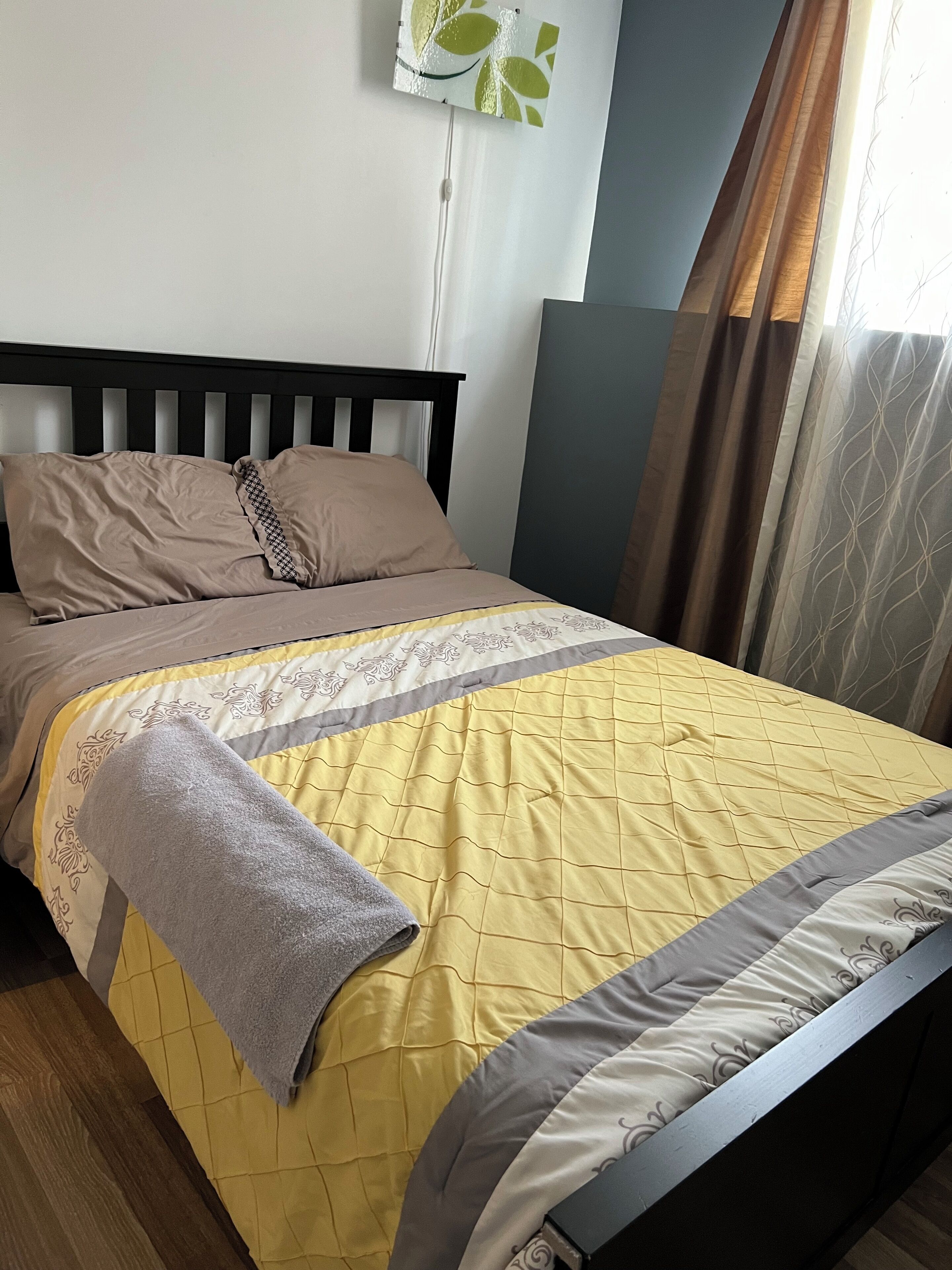 A neatly made bed with a yellow quilt and a grey blanket, a wooden headboard, and a wall-mounted light fixture.