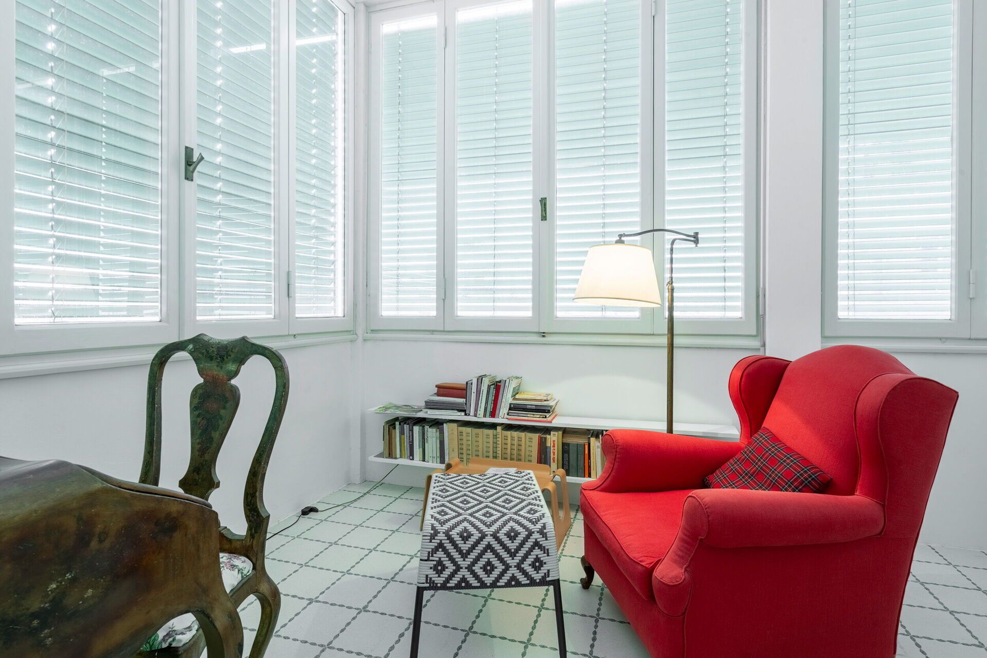 A room with a red armchair, a patterned ottoman, a wooden chair, and a bookshelf filled with books.