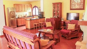 A living room with a sofa, a coffee table, and a TV. There is a kitchen area with wooden cabinets and a refrigerator.