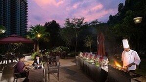 An outdoor dining area with a chef preparing food, tables, chairs, and a building in the background. - TUI Blue The Haven Ipoh (Ipoh)