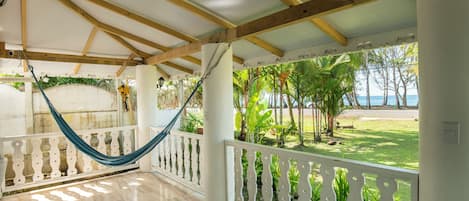 A covered porch with a hammock, white railing, and a view of the beach.