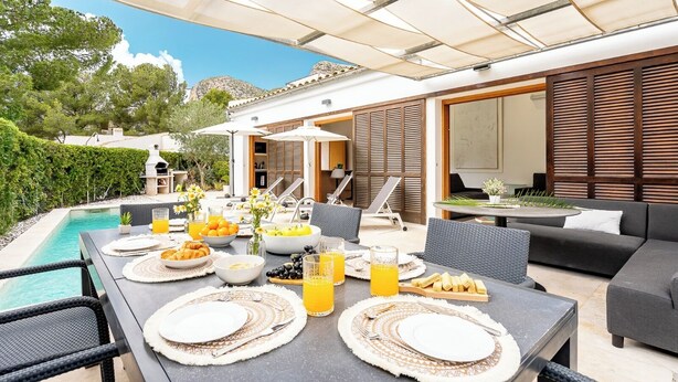 Un espace extérieur aménagé pour les repas, avec une piscine, une table à manger dressée avec des assiettes, des verres et des fruits, entouré de chaises longues et d’un patio couvert.