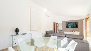 Un salon moderne avec une table à manger en verre, un canapé gris et un téléviseur à écran plat fixé sur un mur recouvert de panneaux en bois.