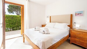 Une chambre avec un lit en bois, du linge de lit blanc, une table de chevet avec une lampe, et une vue sur une haie verdoyante à travers une porte vitrée.