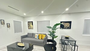 Un salon moderne avec un canapé gris, une table basse noire et un fauteuil noir. On y trouve une plante en pot, une petite table avec un bol, et des œuvres d’art accrochées aux murs.