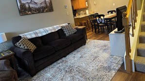 Een woonkamer met een donkerbruine bank, een kamerkleed met een patroon, en een schilderij van bergen.