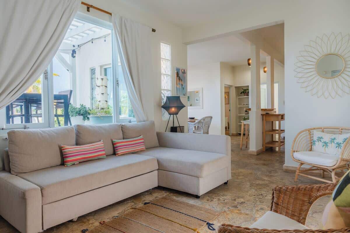 Un salon avec un canapé d’angle beige, un fauteuil en osier et une lampe sur pied.