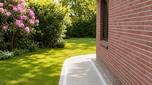 A brick building with a paved pathway leading to a well-maintained lawn and flowering bushes.