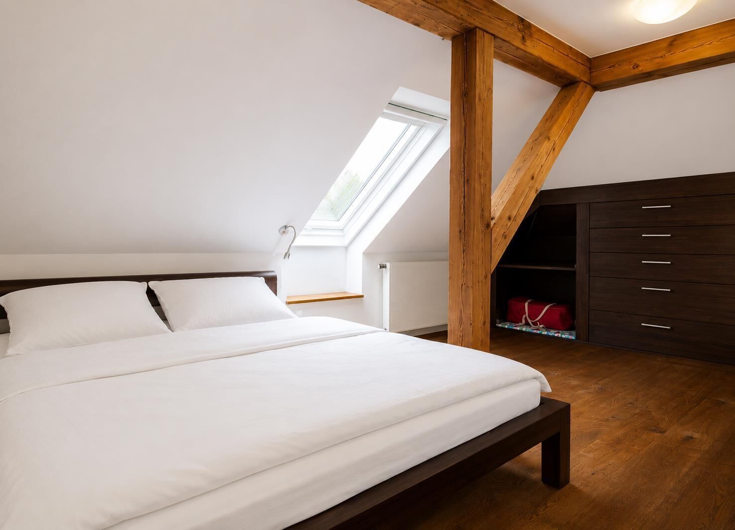 A bedroom with a bed, wooden beams, a skylight, a dresser, and a radiator.