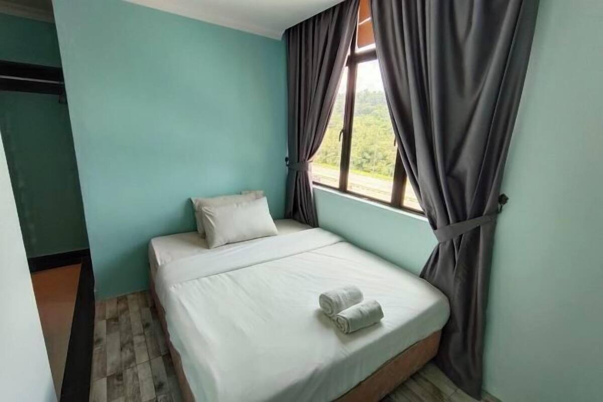 A single bed with white bedding and two rolled towels, a window with curtains, and teal walls.