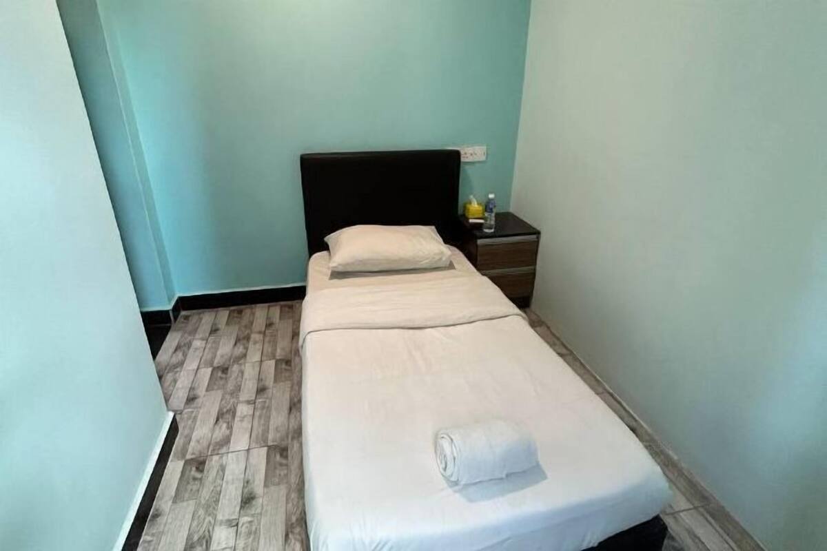 A single bed with a white mattress and pillow, a wooden nightstand with a bottle, and a light blue wall.