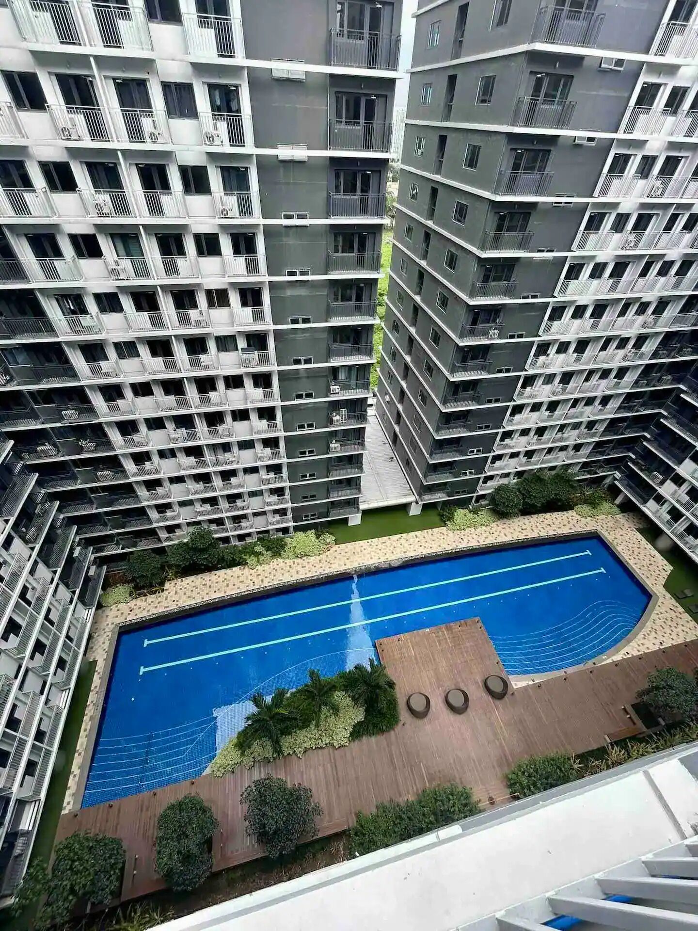 Ein Hochhaus mit Wohneinheiten, mehreren Balkonen, einem Schwimmbad und einer begrünten Anlage mit Bäumen und einer Holzterrasse.