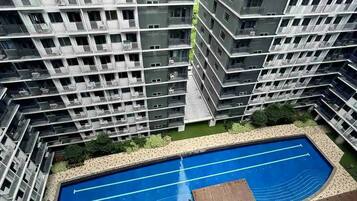 Ein Hochhaus mit Wohneinheiten, mehreren Balkonen, einem Schwimmbad und einer begrünten Anlage mit Bäumen und einer Holzterrasse.