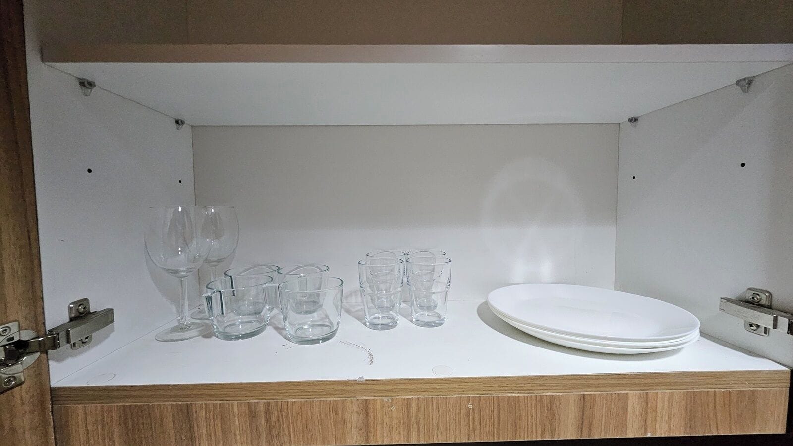 A white cabinet with a wooden base, containing a wine glass, several shot glasses, and a white plate.