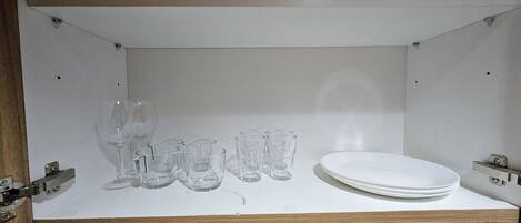 Ett vitt skåp med en träbas, innehållande ett vinglas, flera små glas för starka drycker och en vit tallrik.