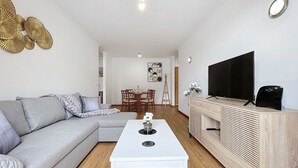 A modern living room with a grey sofa, a white coffee table, a TV on a wooden stand, and a dining area in the background.
