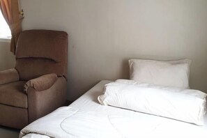 A single bed with white bedding and a brown armchair in a room with plain walls. - Green Homestay (Junrejo)