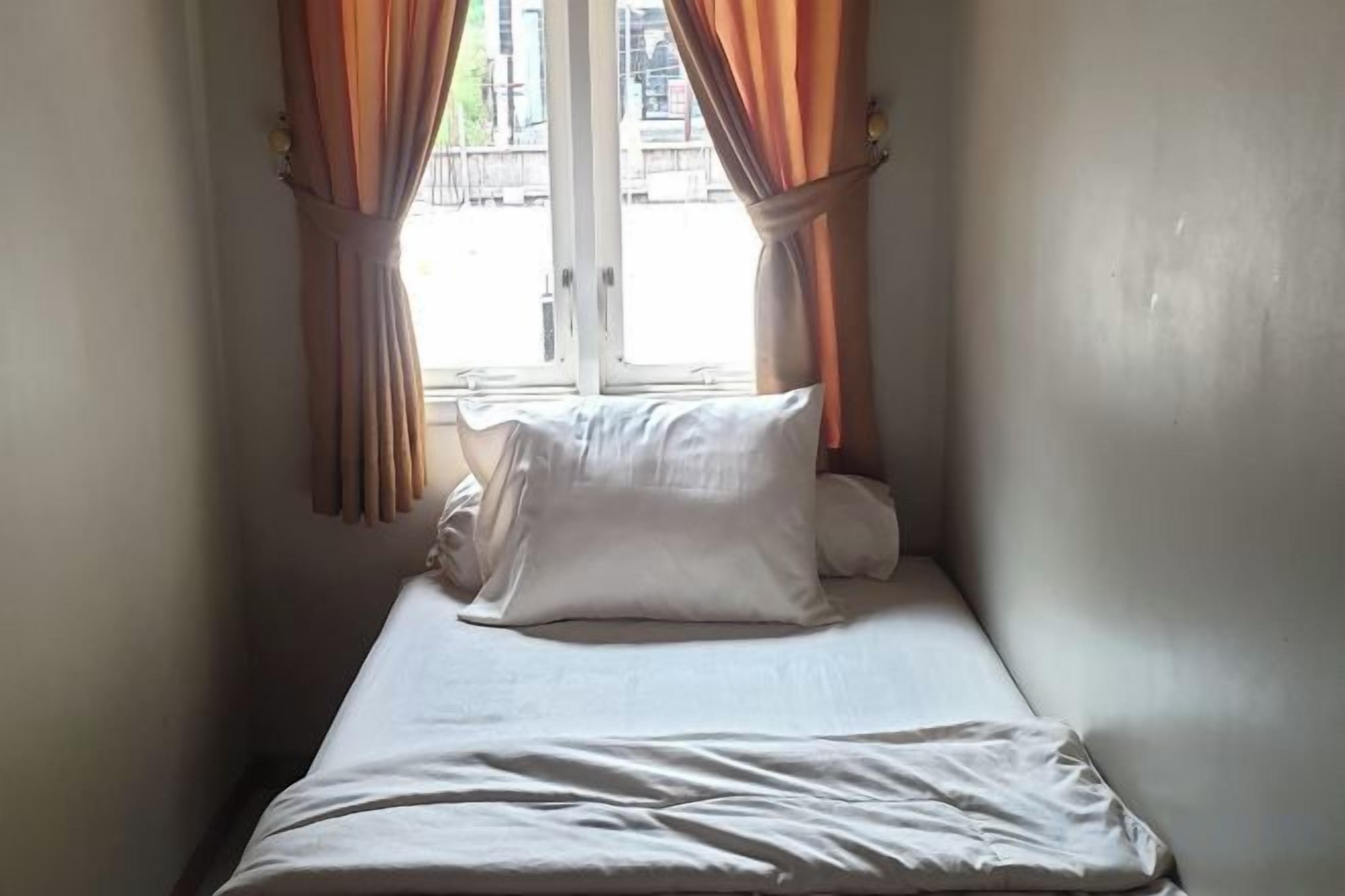 Un lit simple avec une literie blanche, dans une petite pièce dotée d’une fenêtre et de rideaux.