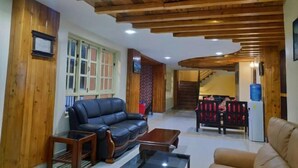 A living room with a wooden ceiling, a black leather sofa, a glass coffee table, and a dining area with red chairs. - Hotel green valley nagarkot (Baluwapati Deupur)