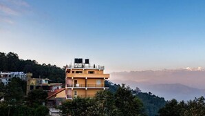 A multi-story building with a flat roof and a balcony, surrounded by trees and other buildings, with a mountainous backdrop. - Hotel green valley nagarkot (Baluwapati Deupur)