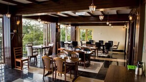 A spacious dining area with multiple tables and chairs, large windows offering views of greenery, and hanging pendant lights. - Hotel green valley nagarkot (Baluwapati Deupur)