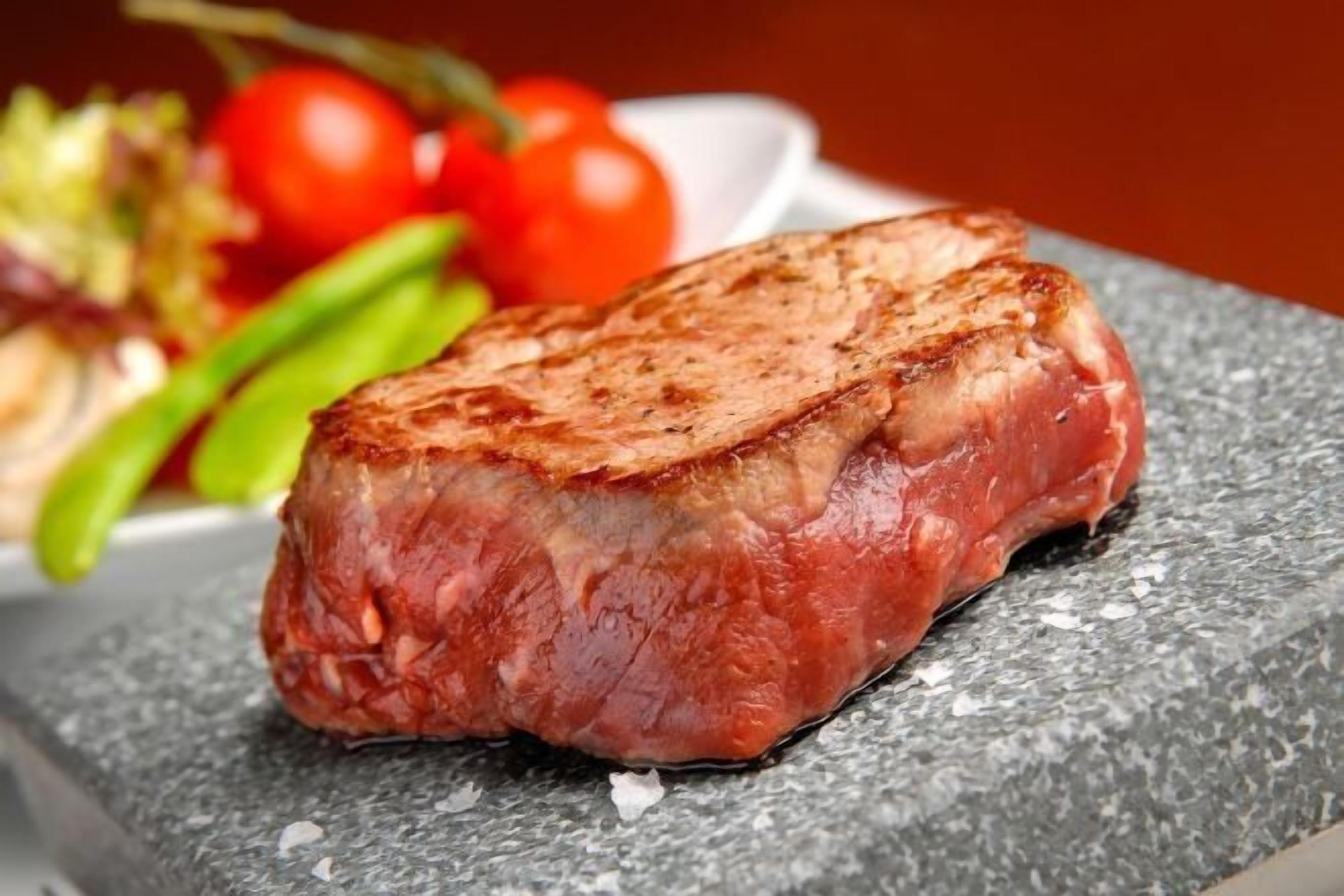A steak on a stone surface with a plate of vegetables in the background.