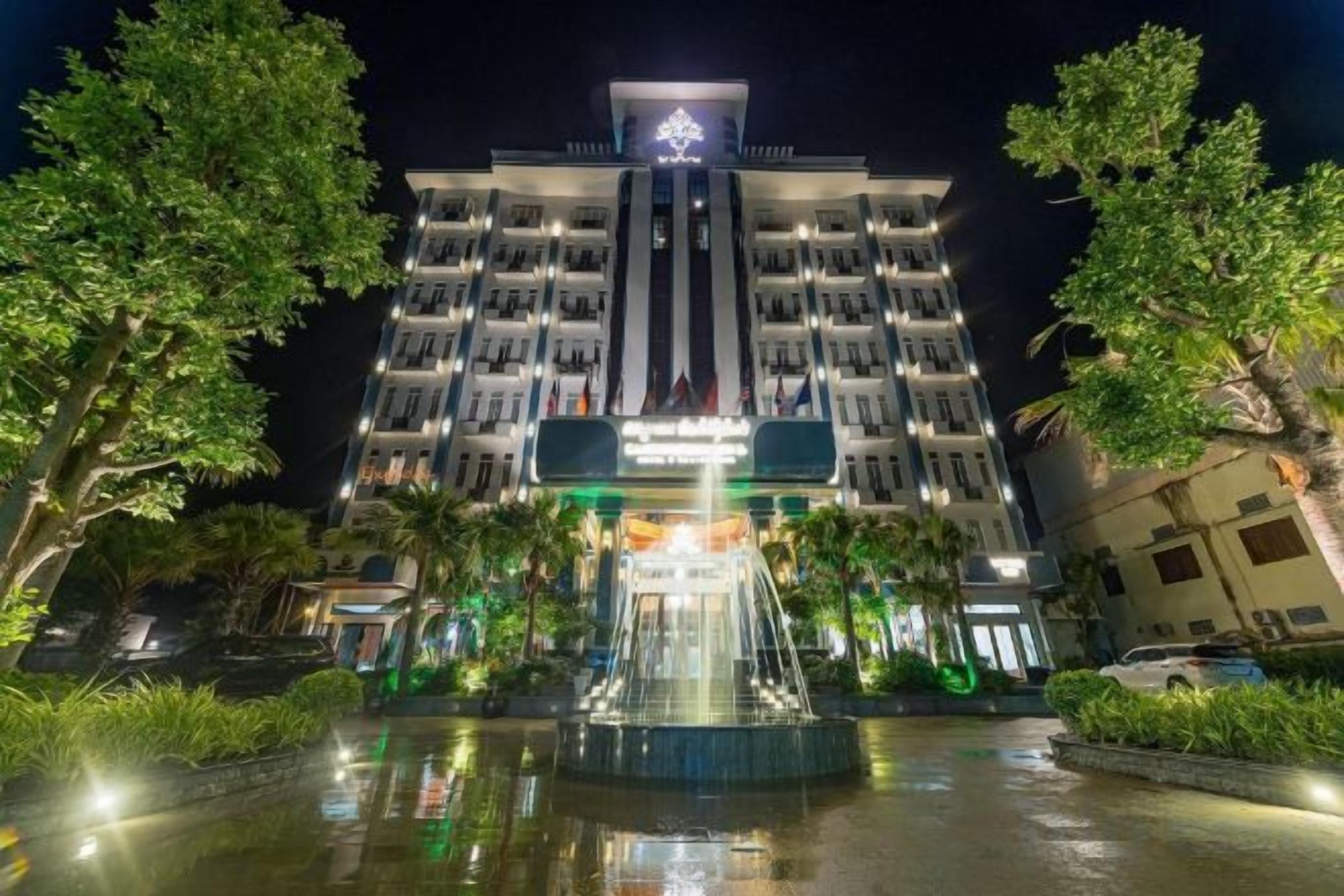 Ein mehrstöckiges Hotel mit einer zentralen Fontäne, umgeben von Grünflächen und beleuchtet mit künstlichem Licht.