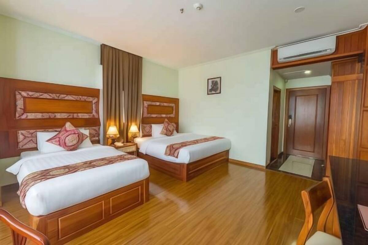 Une chambre d’hôtel avec deux lits, des têtes de lit en bois, une table de chevet avec une lampe, une chaise en bois et une porte donnant sur une salle de bain.