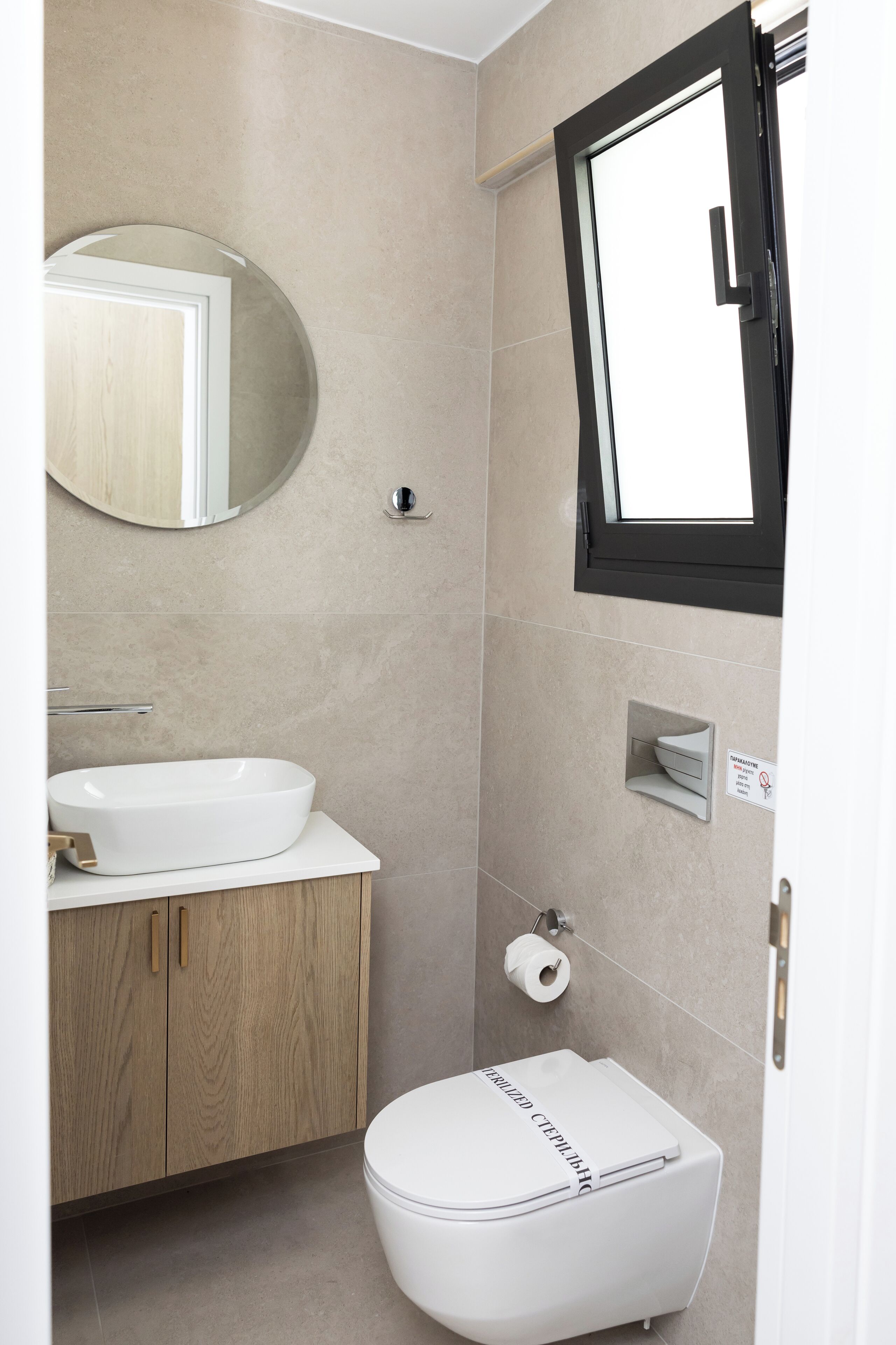 A modern bathroom with a white sink, wooden vanity, a round mirror, a toilet, and a window.