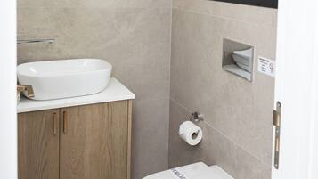 A modern bathroom with a white sink, wooden vanity, a round mirror, a toilet, and a window.