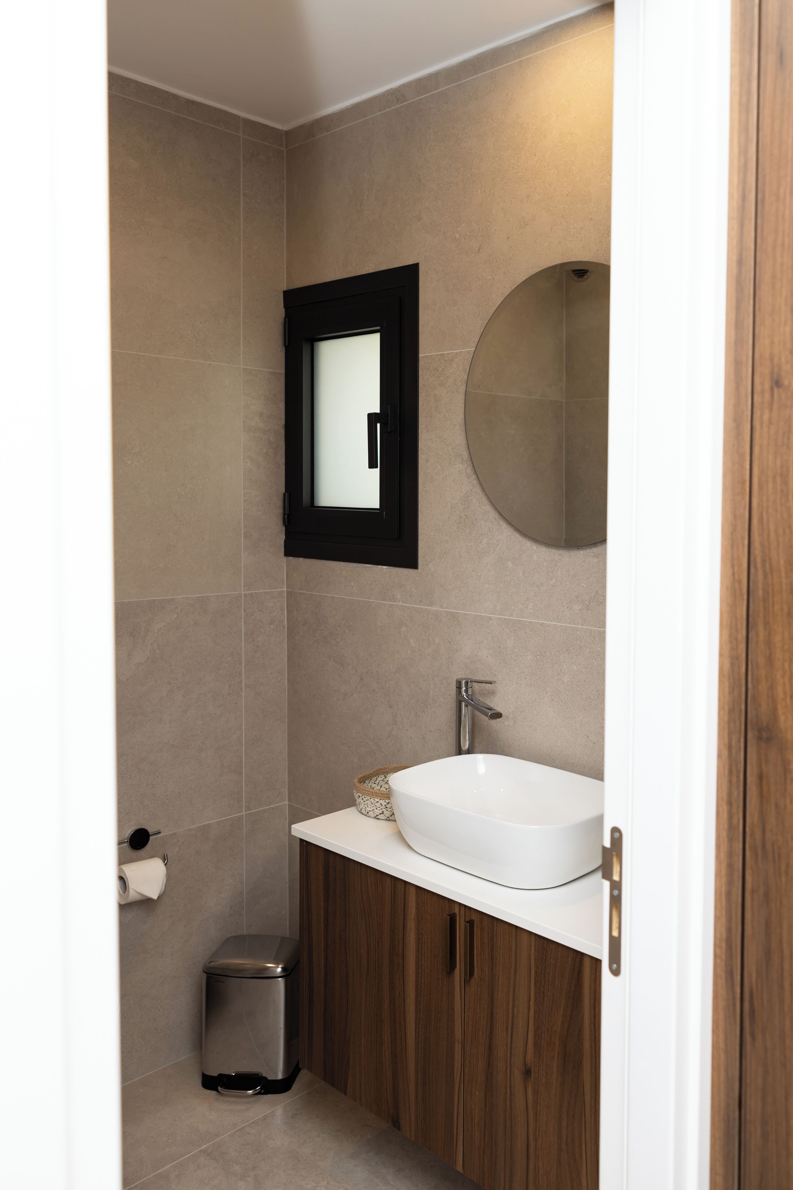 Een badkamer met een witte wastafel, houten kastjes, een ronde spiegel en een kleine prullenbak.