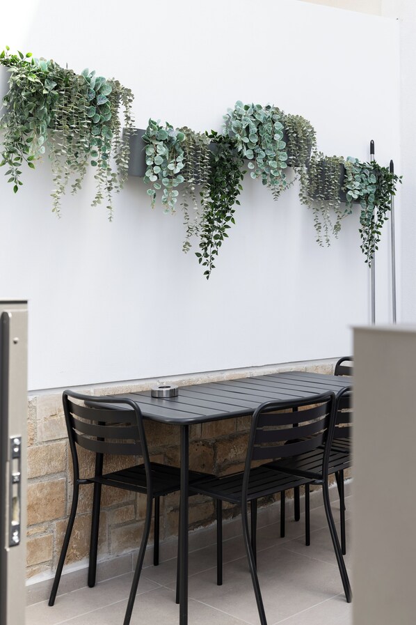 Une terrasse avec une table et des chaises en métal noir, entourée d’un mur blanc orné de plantes vertes suspendues.