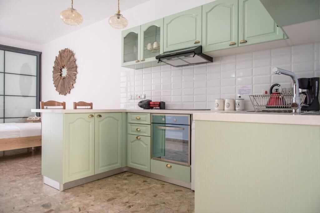 Une cuisine avec des meubles de couleur vert clair, un plan de travail blanc et un four encastré. On y trouve un évier avec un robinet, une machine à café et un miroir décoratif accroché au mur.