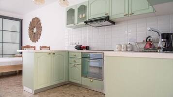Une cuisine avec des meubles de couleur vert clair, un plan de travail blanc et un four encastré. On y trouve un évier avec un robinet, une machine à café et un miroir décoratif accroché au mur.