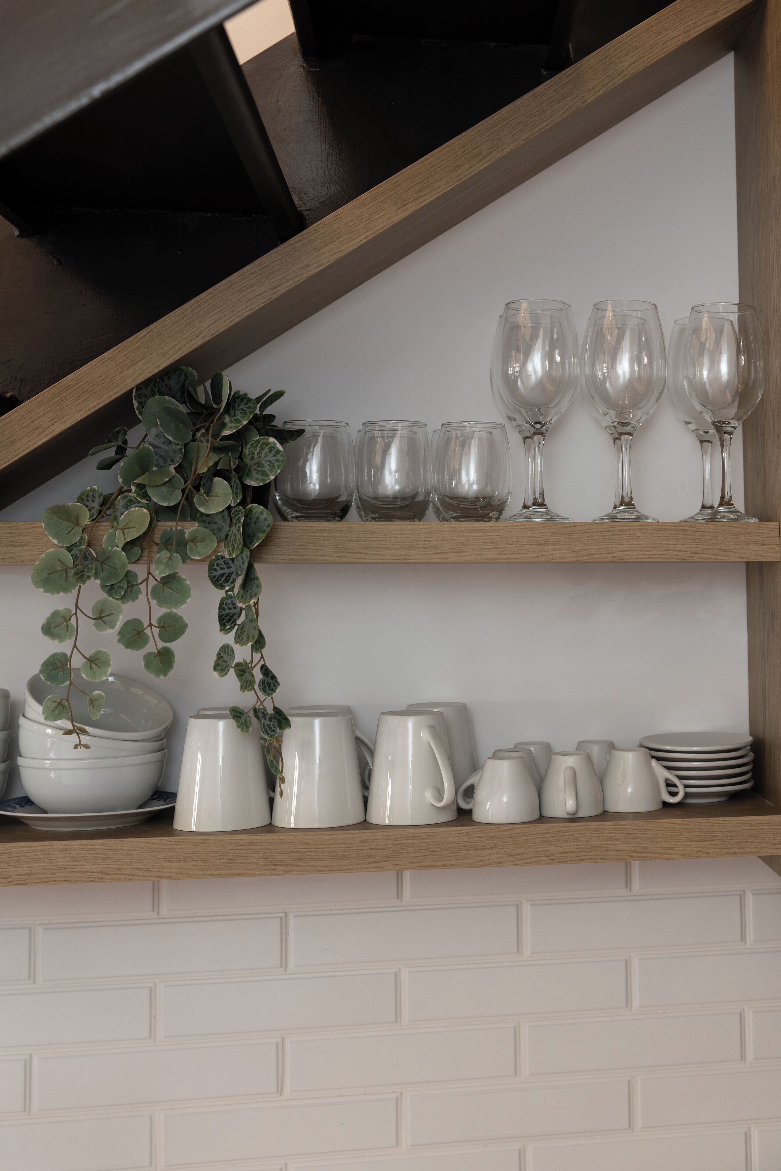 Una cocina con paredes de ladrillo blanco, estantes de madera y varios utensilios de cocina blancos, incluyendo tazas, cuencos y una planta.
