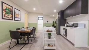 A modern kitchen with white cabinets, a microwave, and a white countertop. There is a dining area with a table and chairs. - Kula Parramatta (MAYS HILL)