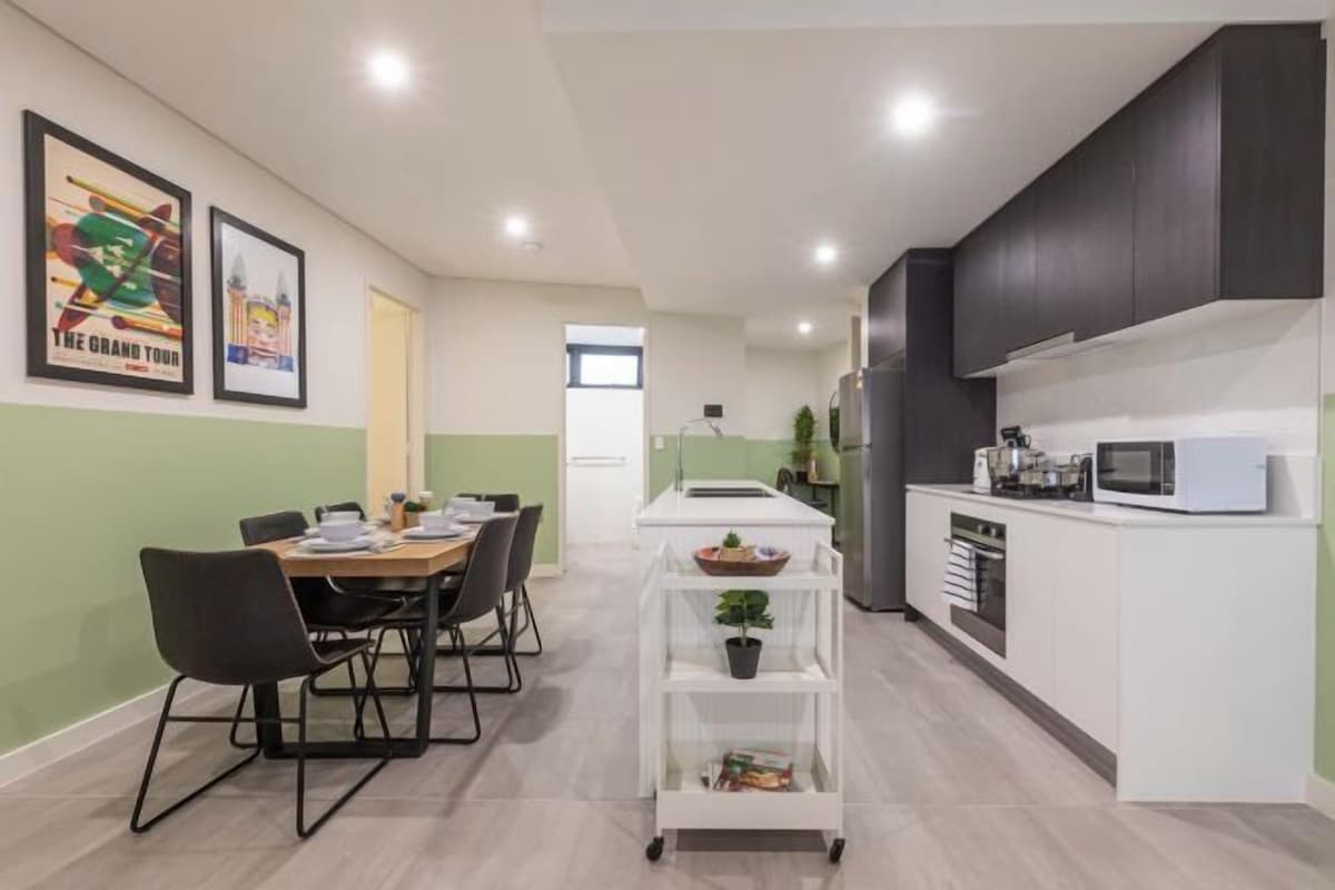 A modern kitchen with white cabinets, a microwave, and a white countertop. There is a dining area with a table and chairs.