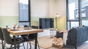 A modern living room with a dining table set for two, a sofa, a TV, and a large window. - Kula Parramatta (MAYS HILL)