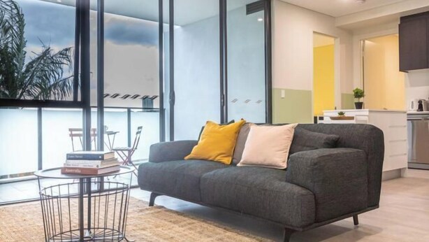 A modern living room with a sofa, a coffee table, and a view through large windows. - Kula Parramatta (MAYS HILL)
