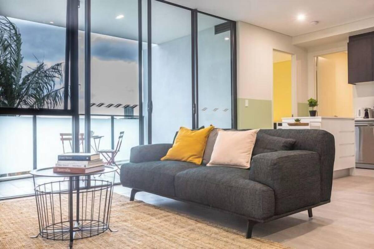 A modern living room with a sofa, a coffee table, and a view through large windows.