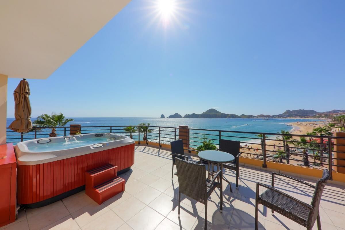 A balcony with a hot tub, chairs, and a table overlooking a beach and ocean.