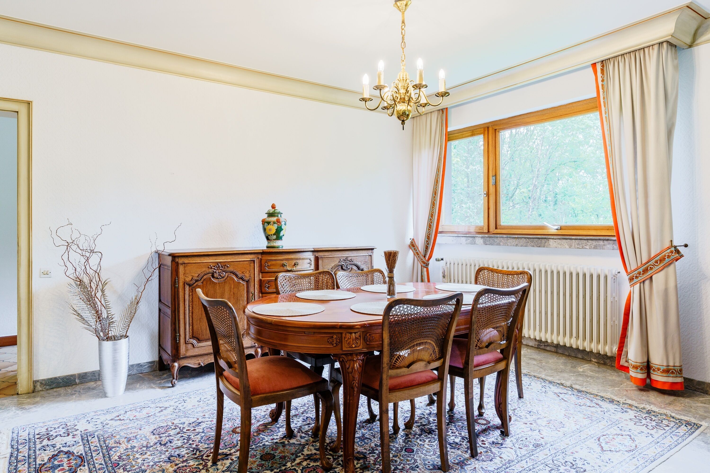 A dining room with a round wooden table, six chairs, a chandelier, a radiator, and a window with curtains.
