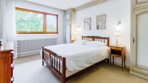 A bedroom with a wooden bed, a window with curtains, a radiator, and two wall-mounted lamps.