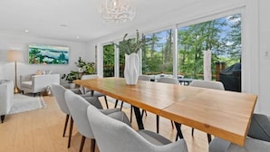 Ruime eethoek met een houten tafel en grijze stoelen, grote ramen met uitzicht op groen, en een moderne kroonluchter.