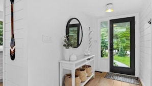 Een schone, witte gang met een spiegel met zwart frame, een witte console tafel met manden, en een deur die uitkomt op een groen buitengebied.